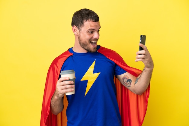 Super Hero Brazilian man isolated on yellow background holding coffee to take away and a mobile