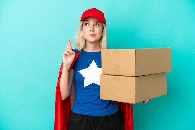 Super Hero caucasian delivery woman isolated on blue background with fingers crossing and wishing the best
