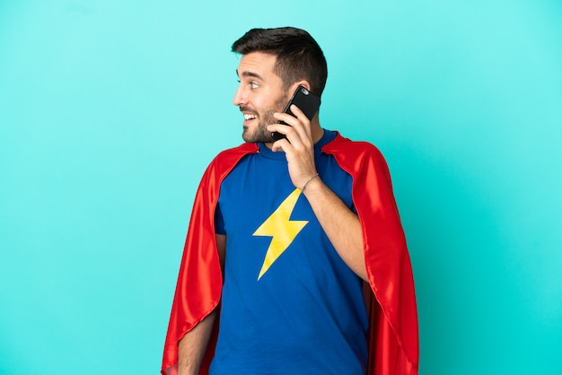 Super Hero caucasian man isolated on blue background keeping a conversation with the mobile phone with someone