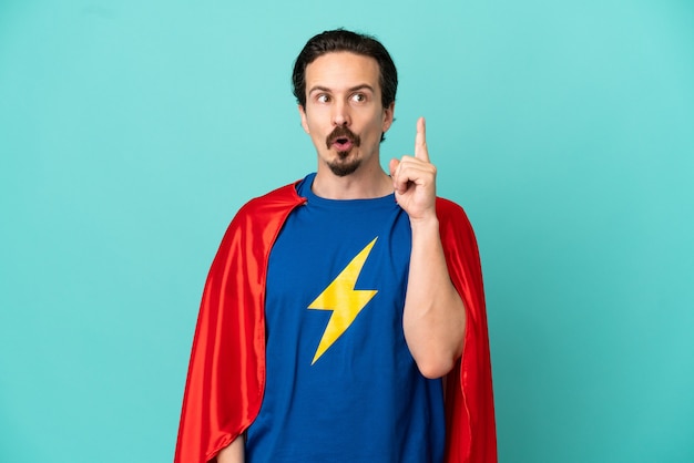 Super Hero caucasian man isolated on blue background thinking an idea pointing the finger up