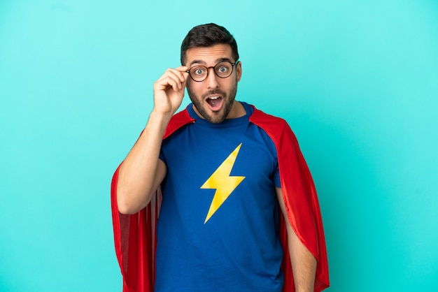 Super Hero caucasian man isolated on blue background with glasses and surprised