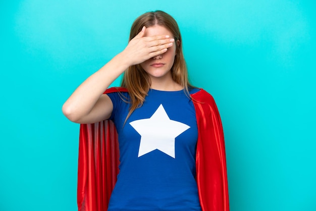 Super hero caucasian woman isolated on blue background covering\
eyes by hands do not want to see something