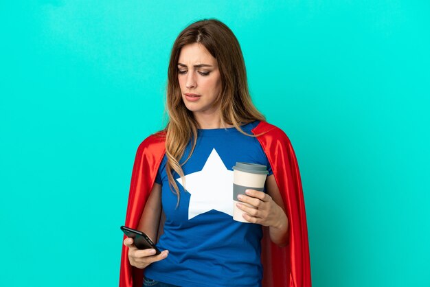 Super Hero caucasian woman isolated on blue background holding coffee to take away and a mobile