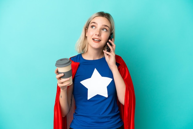 Super Hero caucasian woman isolated on blue background holding coffee to take away and a mobile