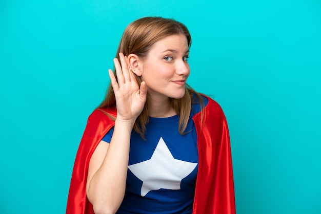 Super Hero caucasian woman isolated on blue background listening to something by putting hand on the ear