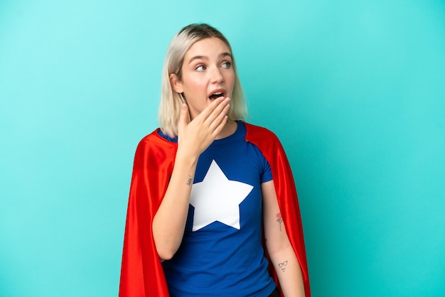 Super Hero caucasian woman isolated on blue background yawning and covering wide open mouth with hand