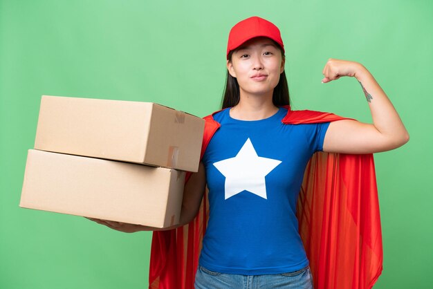Доставка супергероя Азиатская женщина держит коробки на изолированном фоне, делая сильный жест