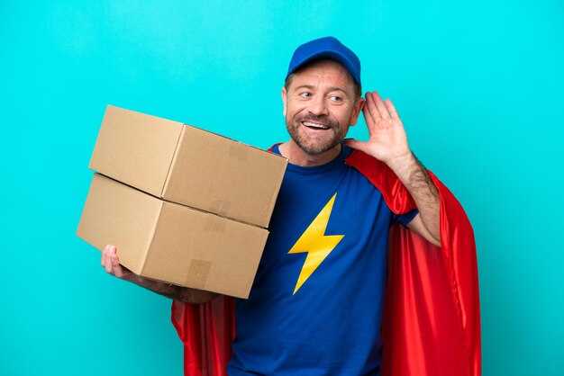 Photo super hero delivery man isolated on blue background listening to something by putting hand on the ear