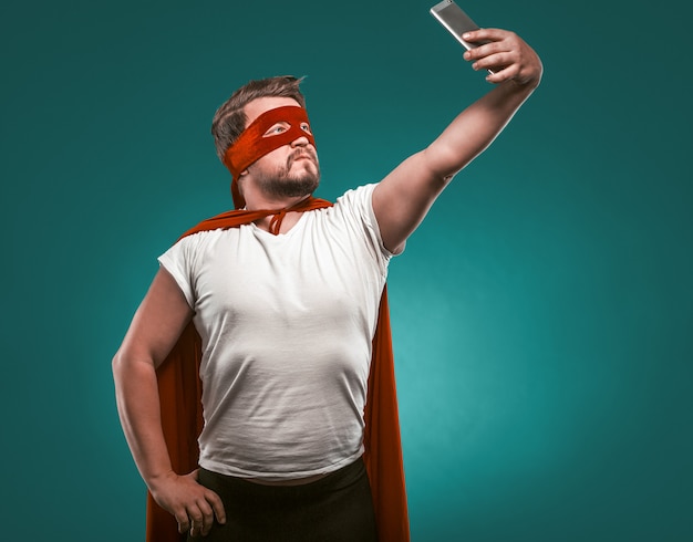 Super Hero man neemt Selfie door mobiele telefoons. Man In Een Super Hero Kostuum In Een Rood Masker En Mantel Geïsoleerd Op Biskaje Groene Achtergrond