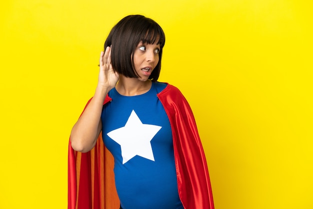 Super Hero pregnant woman isolated on yellow background listening to something by putting hand on the ear