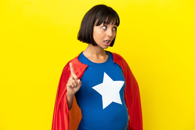 Super Hero pregnant woman isolated on yellow background thinking an idea pointing the finger up
