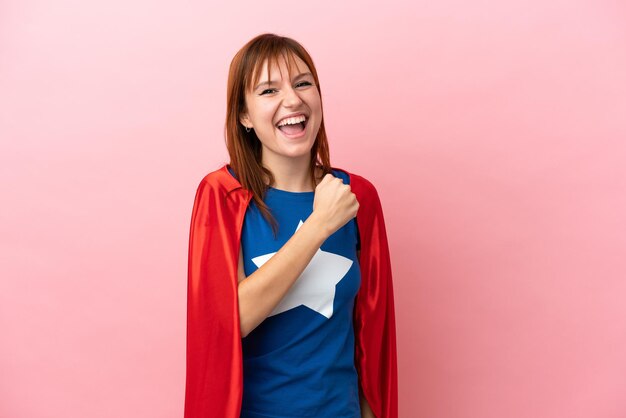 Super Hero redhead girl isolated on pink background celebrating a victory