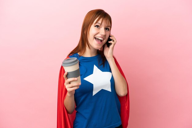 Super Hero redhead girl isolated on pink background holding coffee to take away and a mobile