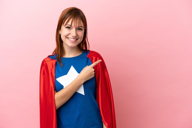 Super Hero redhead girl isolated on pink background pointing to the side to present a product