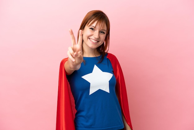 Super Hero redhead girl isolated on pink background smiling and showing victory sign