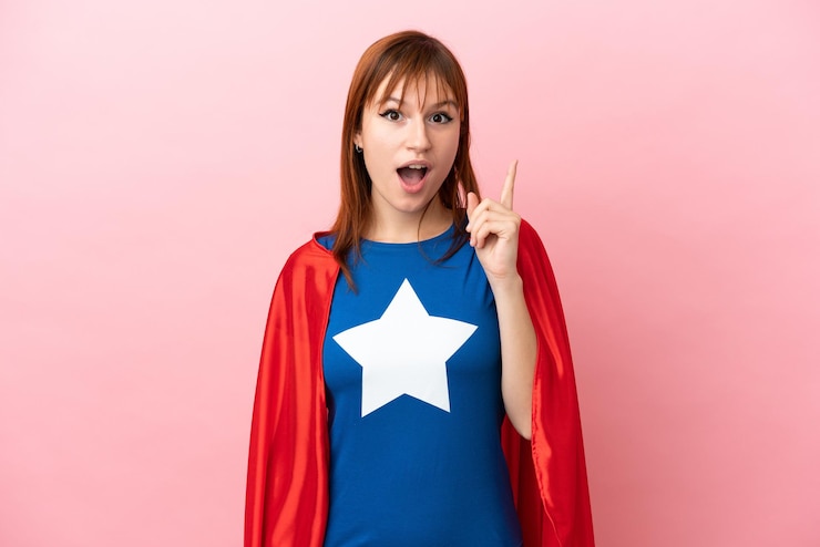 Photo super hero redhead girl isolated on pink background thinking an idea pointing the finger up
