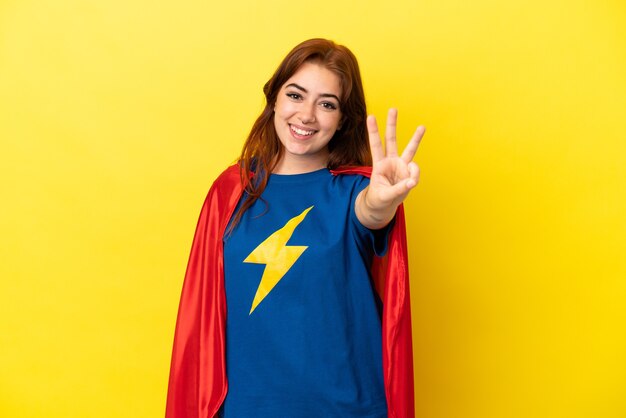 Super Hero redhead woman isolated on yellow background happy and counting three with fingers