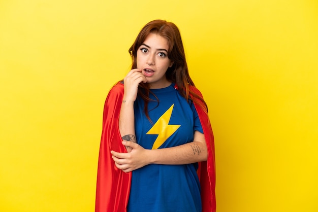 Super Hero redhead woman isolated on yellow background surprised and shocked while looking right