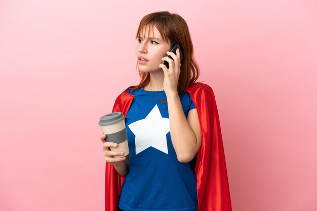 Super Hero roodharig meisje geïsoleerd op roze achtergrond met koffie om mee te nemen en een mobiel