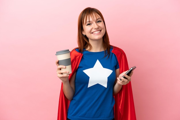 Super Hero roodharig meisje geïsoleerd op roze achtergrond met koffie om mee te nemen en een mobiel