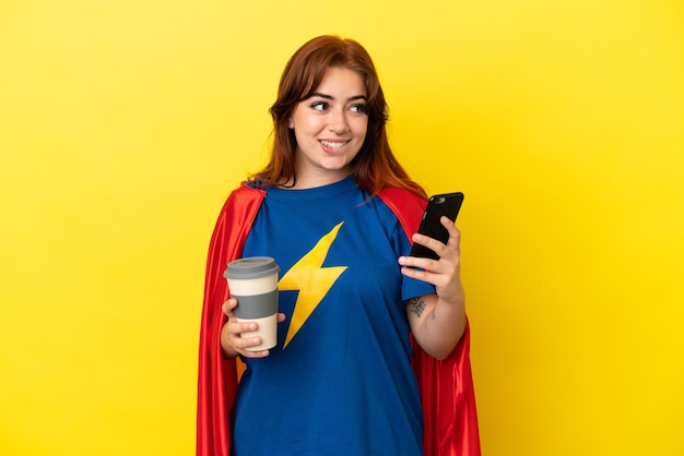 Super Hero roodharige vrouw geïsoleerd op gele achtergrond met koffie om mee te nemen en een mobiel terwijl ze iets denkt