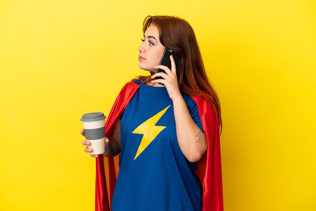 Super Hero roodharige vrouw geïsoleerd op gele achtergrond met koffie om mee te nemen en een mobiel