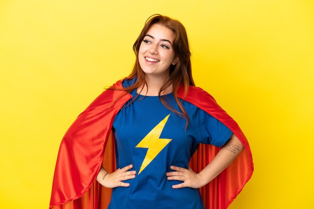 Super Hero roodharige vrouw geïsoleerd op gele achtergrond poseren met armen op heup en lachend