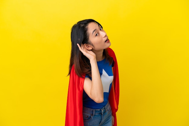 Super hero Vietnamese woman isolated  on yellow background listening to something by putting hand on the ear