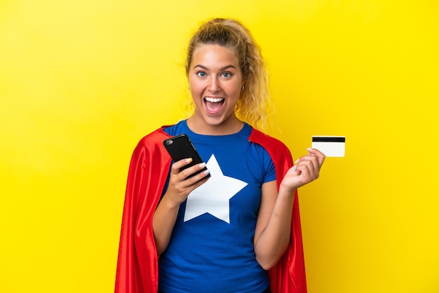 Super Hero woman isolated on yellow background buying with the mobile and holding a credit card with surprised expression