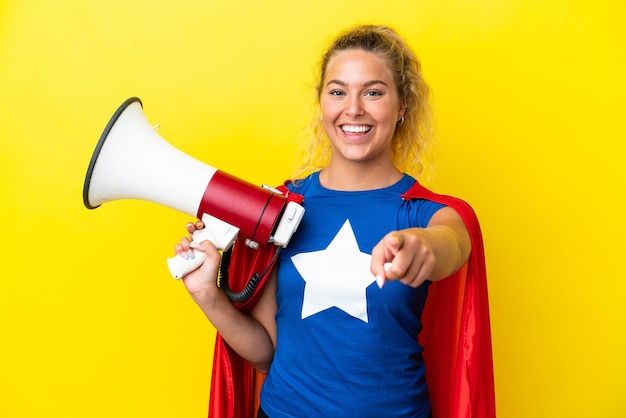 Super Hero woman isolated on yellow background holding a megaphone and smiling while pointing to the front