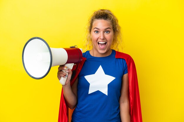 Super Hero woman isolated on yellow background holding a megaphone and with surprise expression