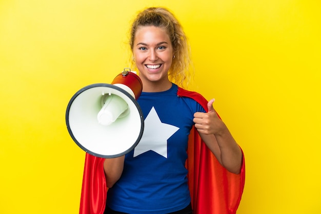 Super Hero woman isolated on yellow background holding a megaphone with thumb up