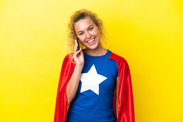 Super Hero woman isolated on yellow background keeping a conversation with the mobile phone with someone