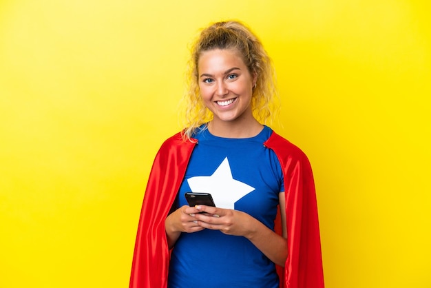 Super Hero woman isolated on yellow background looking at the camera and smiling while using the mobile