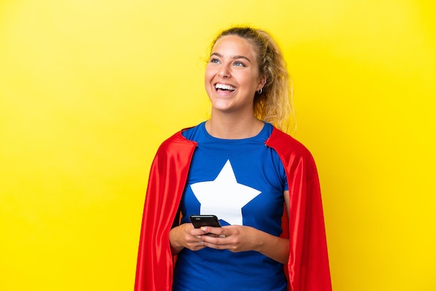 Super hero woman isolated on yellow background using mobile phone and looking up