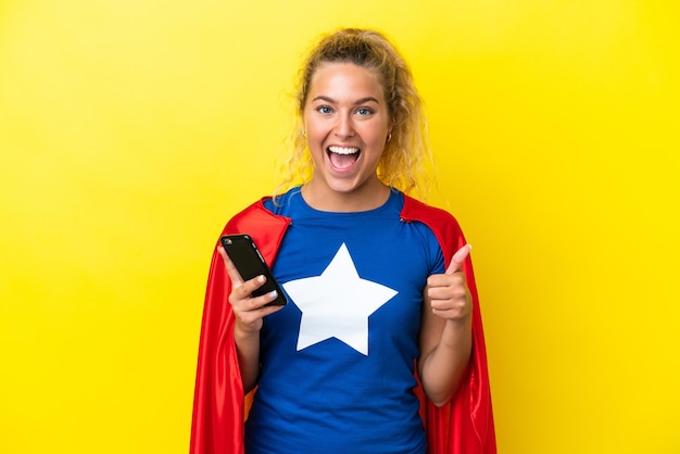 Super Hero woman isolated on yellow background using mobile phone while doing thumbs up