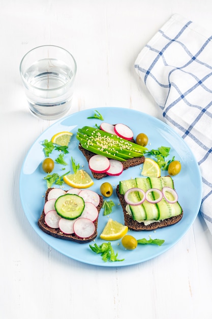 Superfood open vegetarian sandwich with different toppings: avocado, cucumber, radish on plate and glass of water on white background. Healthy eating. Organic and veggie food