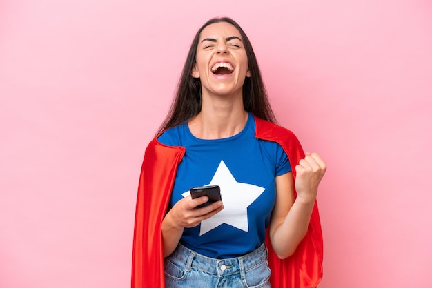 Superheld blanke vrouw geïsoleerd op roze achtergrond met telefoon in overwinningspositie