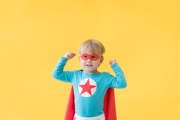 黄色い紙の背景にスーパーヒーローの子。赤いマスクとマントを身に着けているスーパーヒーローの子供。子供の頃の夢と想像力の概念