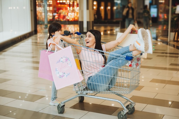スーパーマーケットでは、娘がカートに座っている女性と一緒にカートを押します 幸せな家族が店のカートに元気に駆けつけます