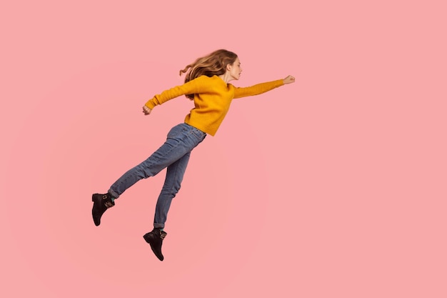超大国。セーターとデニムで勝利に向かって空を飛んでいる決心した真面目な生姜の女の子の肖像画、スーパーヒーローは自由にそして自信を持って目標を達成します。屋内スタジオはピンクの背景を撮影しました