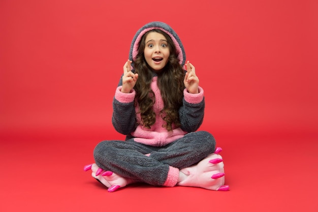 Photo superstitious little girl in easter bunny pajamas make wish keeping fingers crossed for luck red background superstition