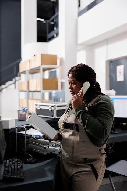 Supervisor looking at white cardboard box while talking with remote customer , discussing online order details using landline phone. African american employee working at merchandise inventory