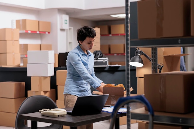 Supervisor putting trendy blouse in cardboard boxes preparing\
customers orders for delivery, checking shipping detalies on laptop\
computer in storehouse, worker analyzing transportation\
logistics