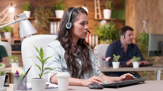 Support customer woman with operator headphone sitting at desk in front of computer communicating with call center customers of telemarketing service in startup office. Helpline concept