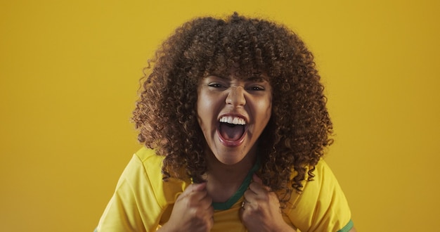 Supporter van Brazilië. Braziliaanse krullend haar vrouw fan vieren op voetbal, voetbalwedstrijd op gele achtergrond. Brazilië kleuren.