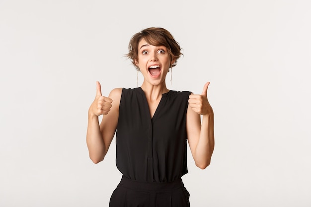 Supportive young woman showing thumbs-up, making compliment. Girl praising great work, standing white amazed.