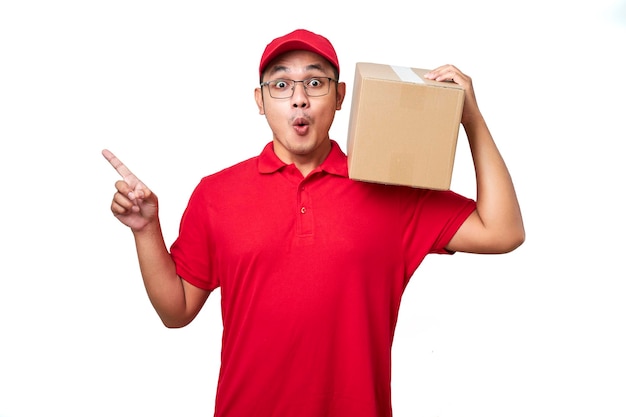 Удивленный азиатский курьер в красной рубашке и кепке держит коробку на плече и указывает влево