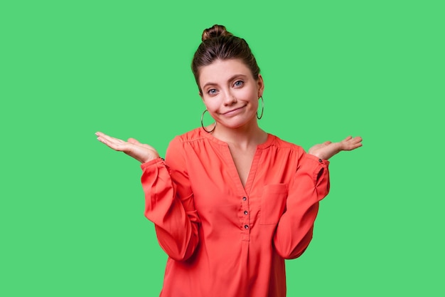 Not sure Portrait of uncertain confused woman with bun hairstyle big earrings and in red blouse shrugging shoulders showing i don39t know gesture indoor studio shot isolated on green background