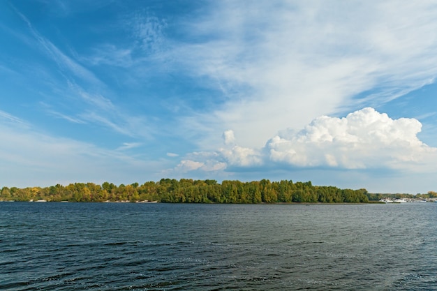 Surface of the river with views of the beach covered with a variety of trees under the massive clouds.